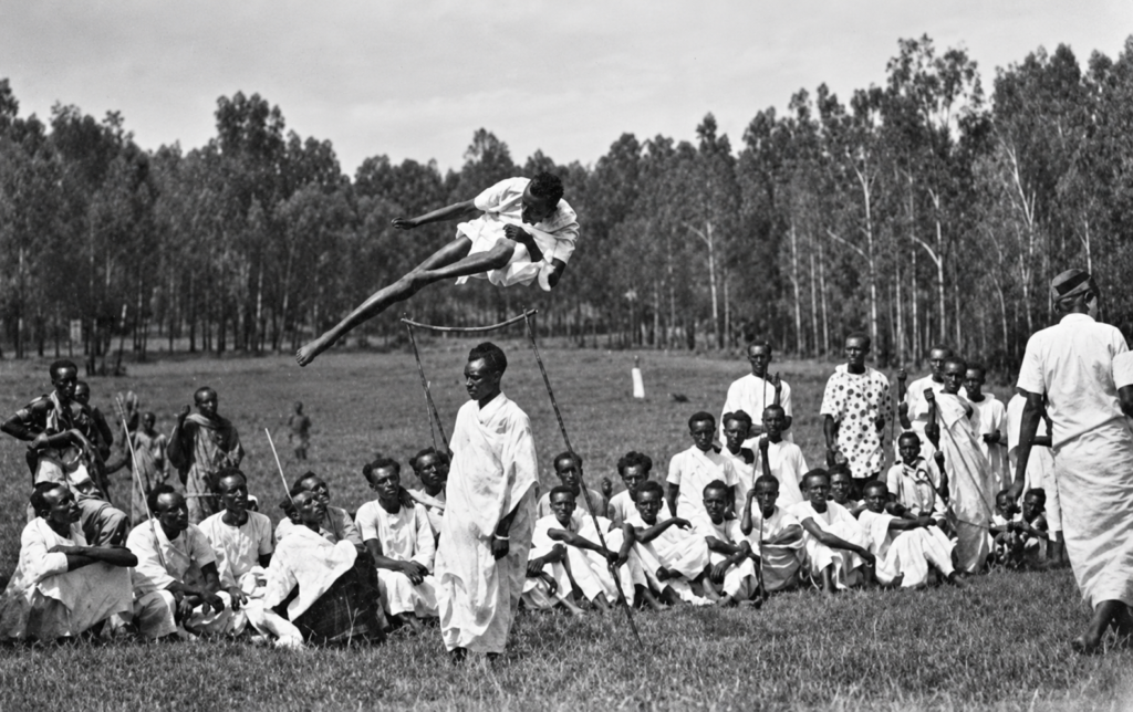 Black and white photograph of an athlete clearing a high jump as a gathered community watches.