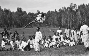 Black and white photograph of an athlete clearing a high jump as a gathered community watches.