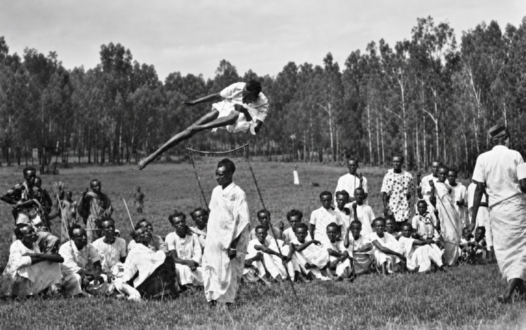 Black and white photograph of an athlete clearing a high jump as a gathered community watches.