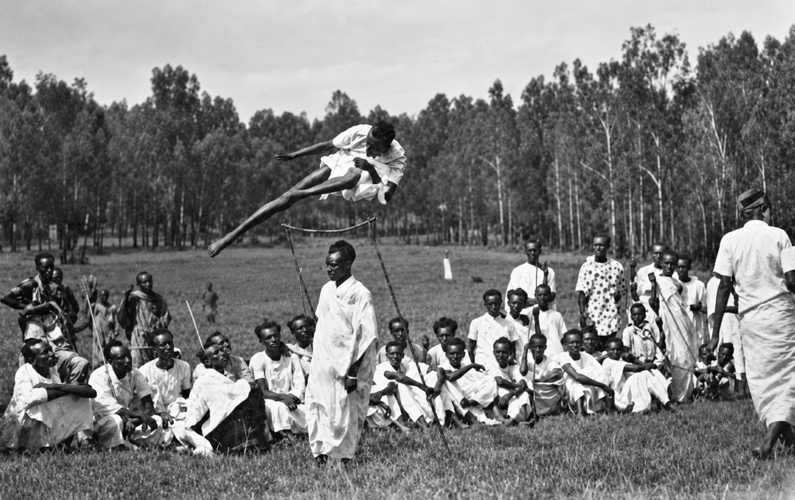 Black and white photograph of an athlete clearing a high jump as a gathered community watches.